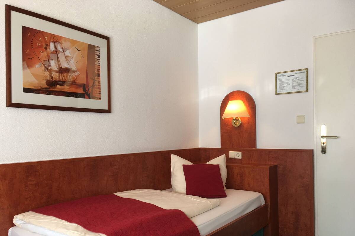 Una cama individual con cabecera de madera, una manta roja y un cuadro con un barco de vela en la pared.