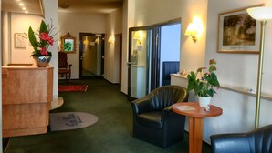 Vestíbulo de hotel con mostrador de recepción, un sillón de cuero, una mesita con un jarrón de flores y un cuadro en la pared.