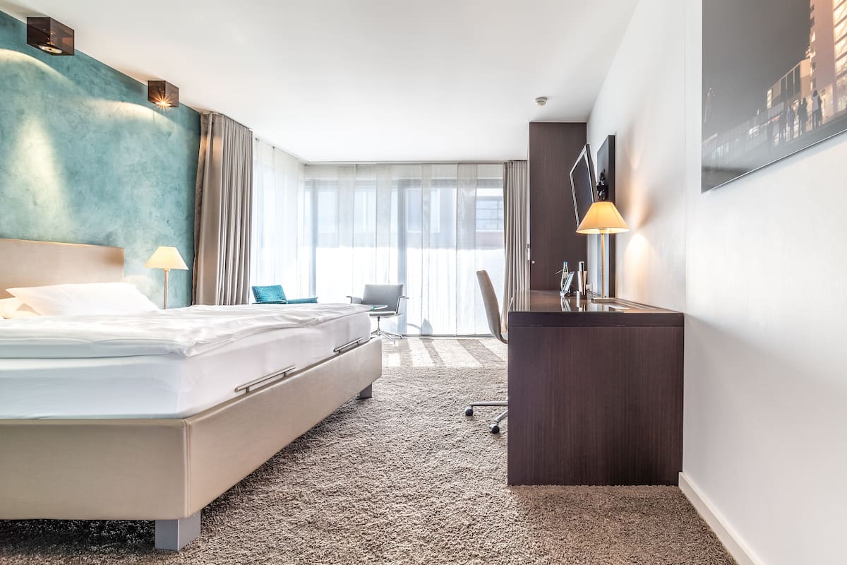 Habitación de hotel con una cama grande, un escritorio, una silla y vistas a la ciudad desde la ventana.
