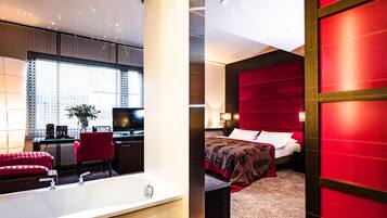 Ein modernes Hotelzimmer mit einer großzügigen Badewanne, einem Schreibtisch mit Computer und einem bequemen Bett mit roter Kopfteil.