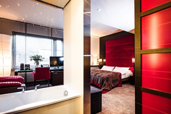 A modern hotel room with a large bathtub, a desk with a computer, and a comfortable bed with a red headboard.