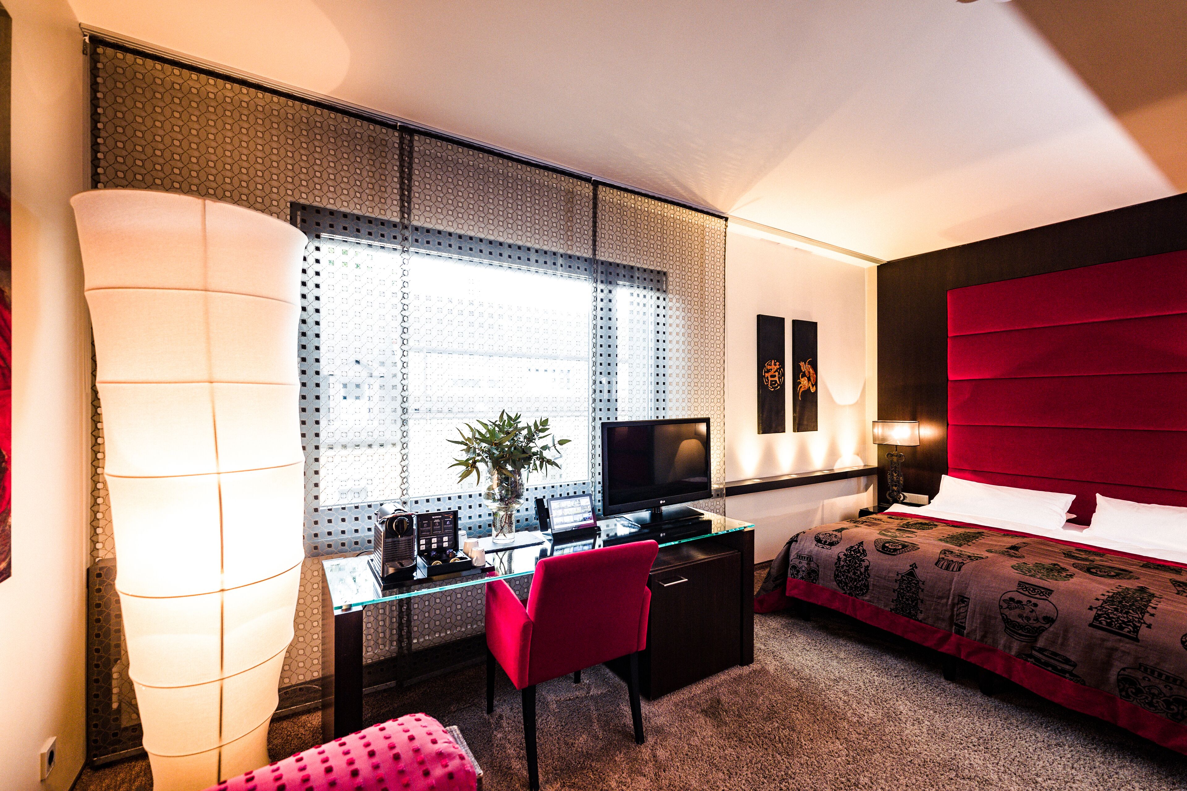 A modern hotel room with a glass desk, a red chair, a bed with a red headboard, and a large window with patterned blinds.