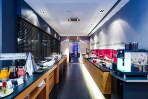 Un hall d’hôtel moderne doté d’un long comptoir d’accueil, d’un espace dédié aux boissons chaudes et d’un buffet proposant une variété de plats et de boissons.