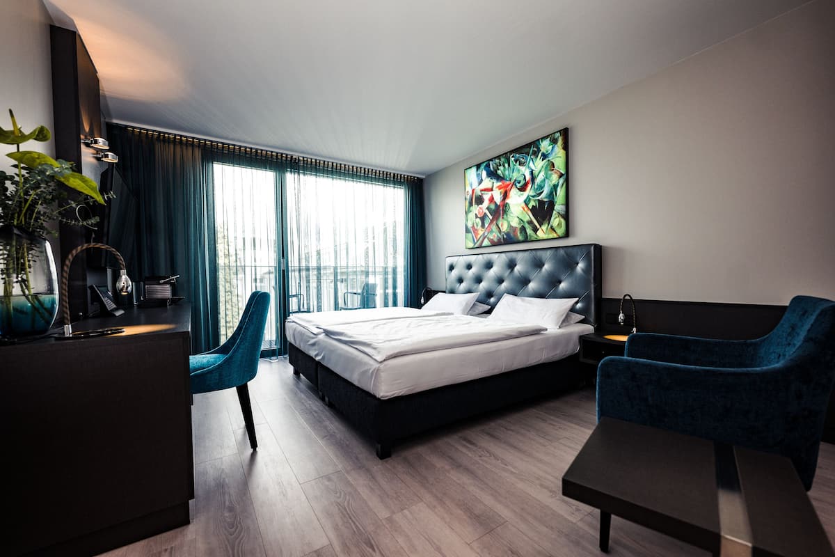 Una habitación de hotel moderna con una cama grande, un escritorio y una silla. Hay una ventana con cortinas y un cuadro en la pared.