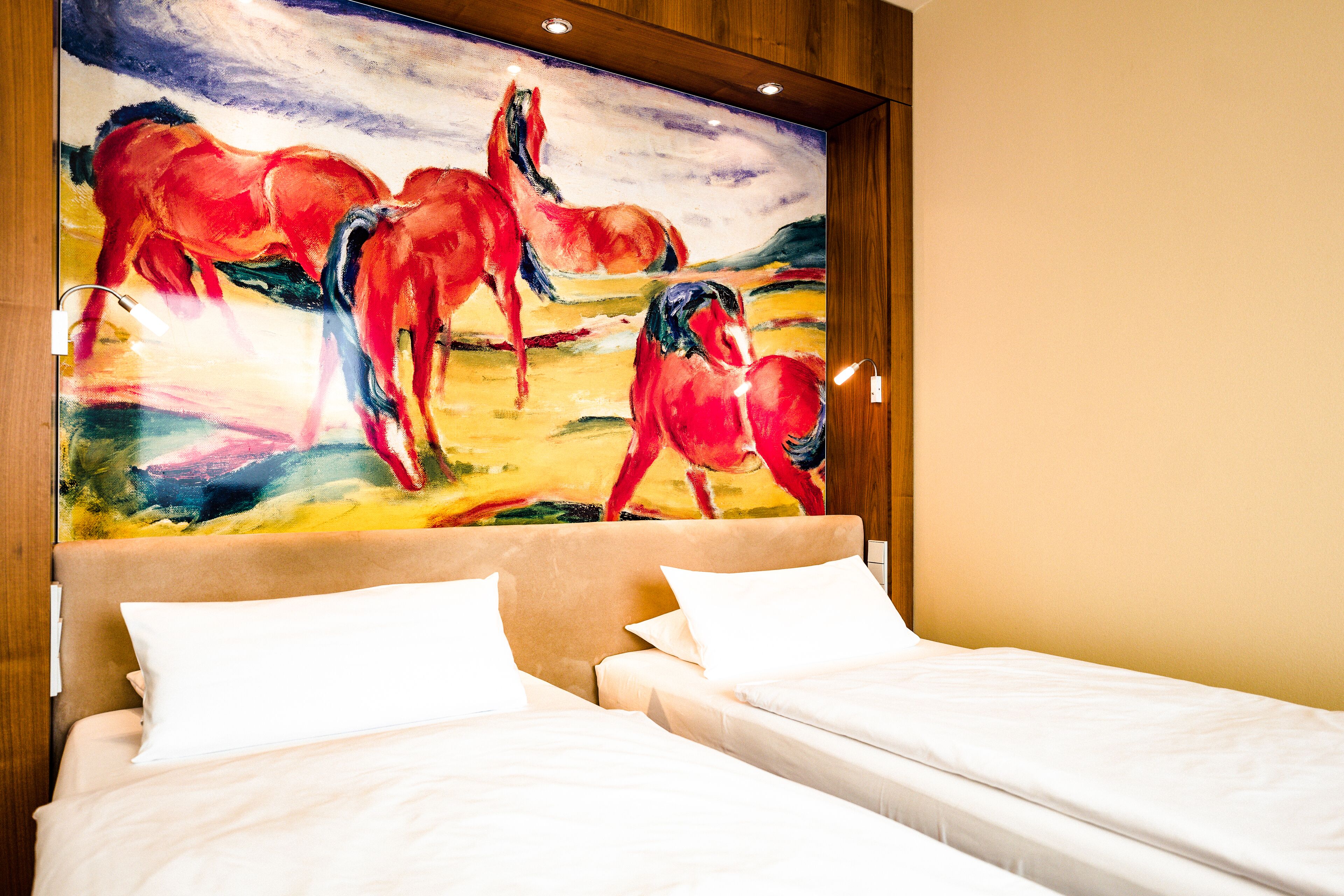 A hotel room with a large painting of horses on the wall, two beds with white linens, and a wooden headboard.