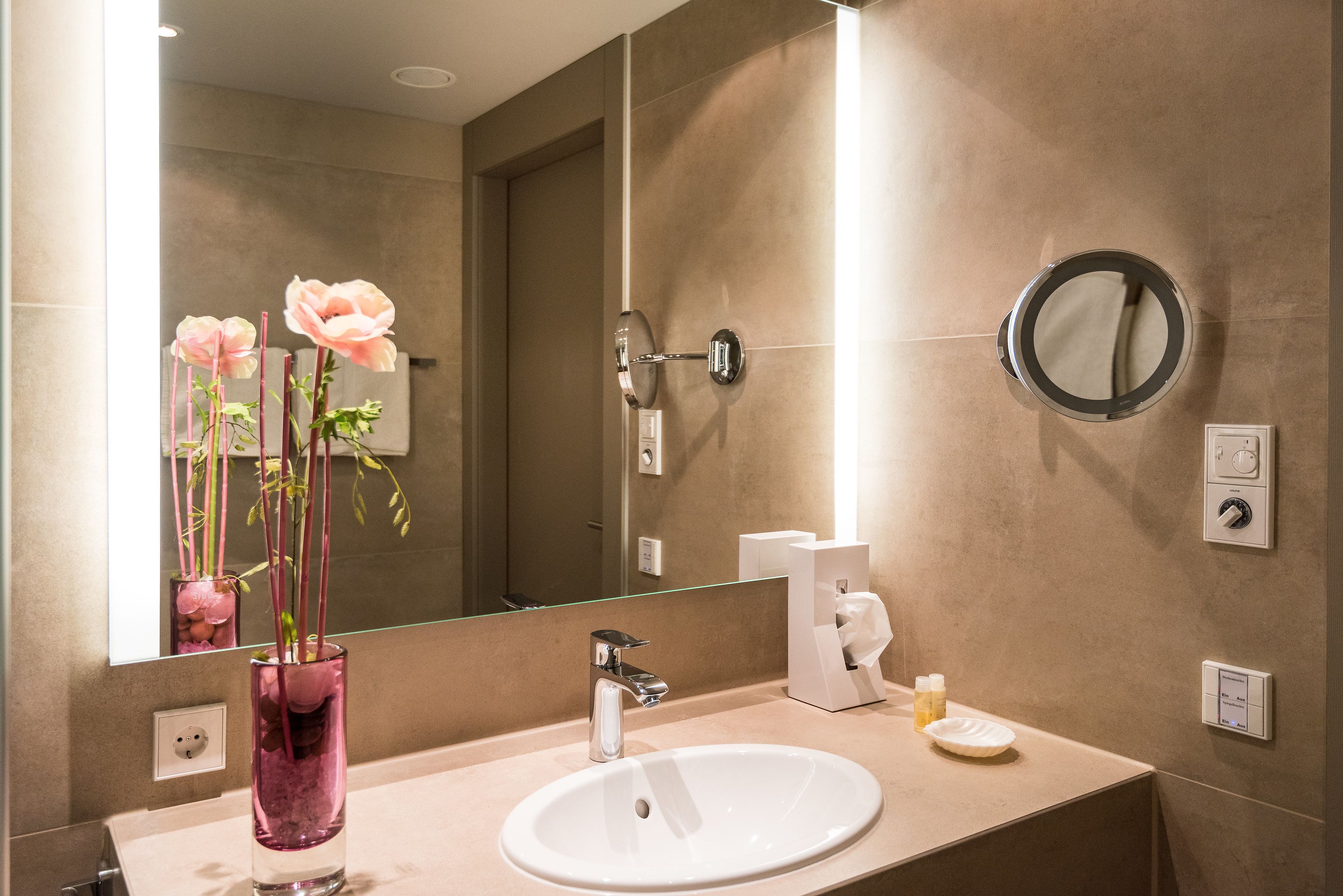 Une salle de bain avec un grand miroir, un lavabo, un distributeur de serviettes et un vase rempli de fleurs.