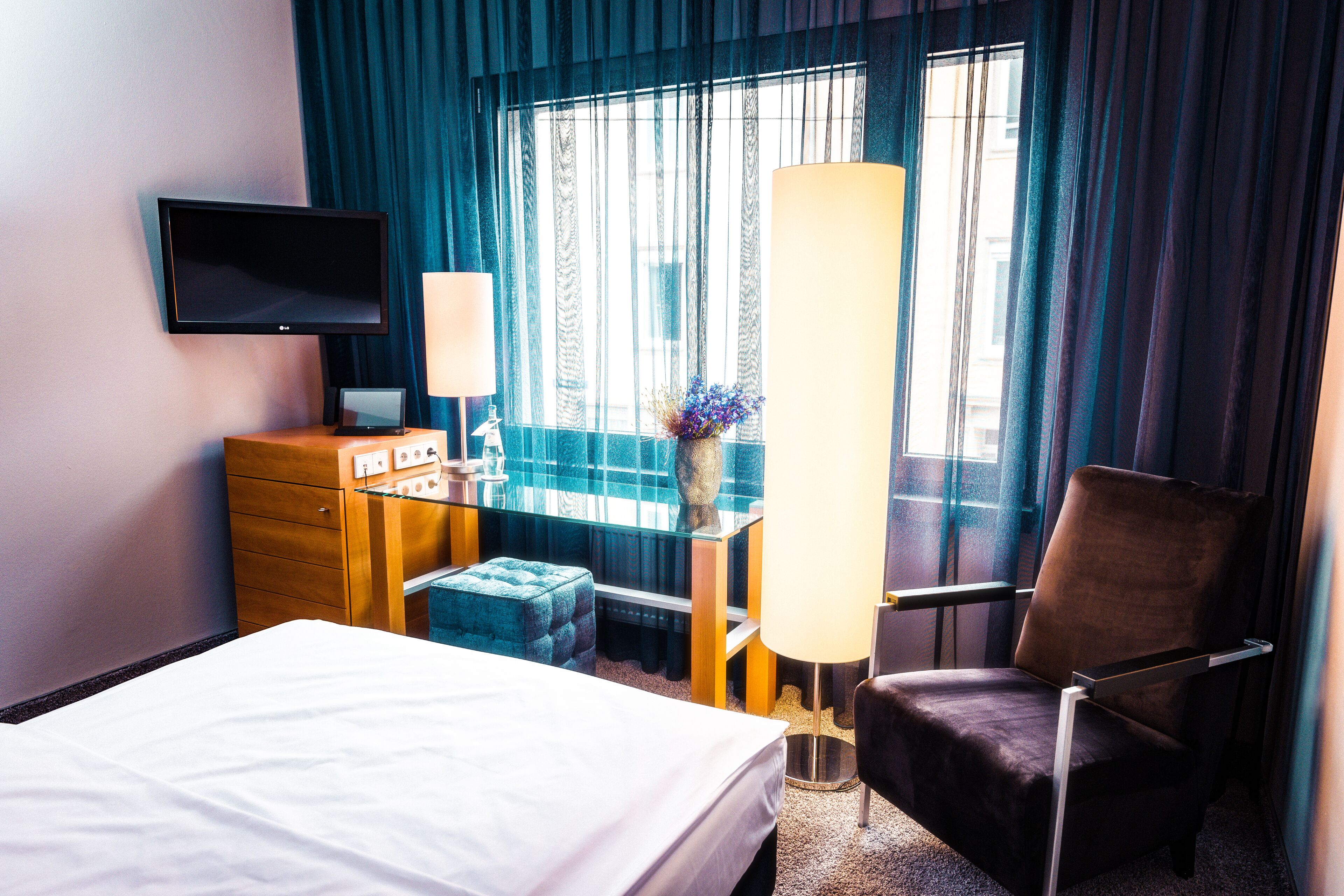 Habitación de hotel con cama, escritorio con lámpara, silla, televisión y ventana con cortinas.