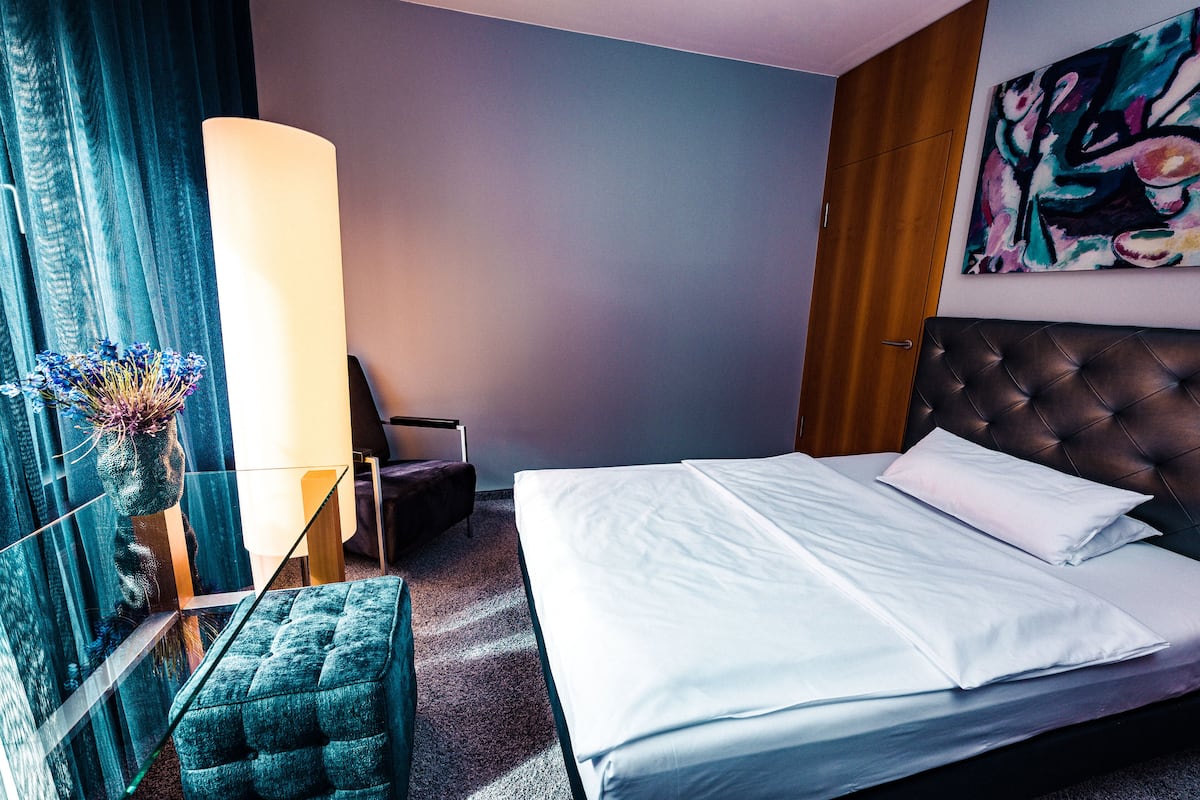 Una habitación de hotel con una cama grande, un cabecero capitonado, una mesita de noche, una silla, un florero con flores y un cuadro en la pared.