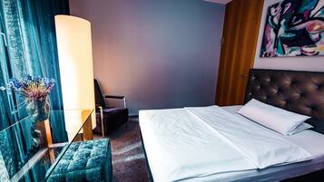 A hotel room with a large bed, a tufted headboard, a bedside table, a chair, a vase with flowers, and a painting on the wall.