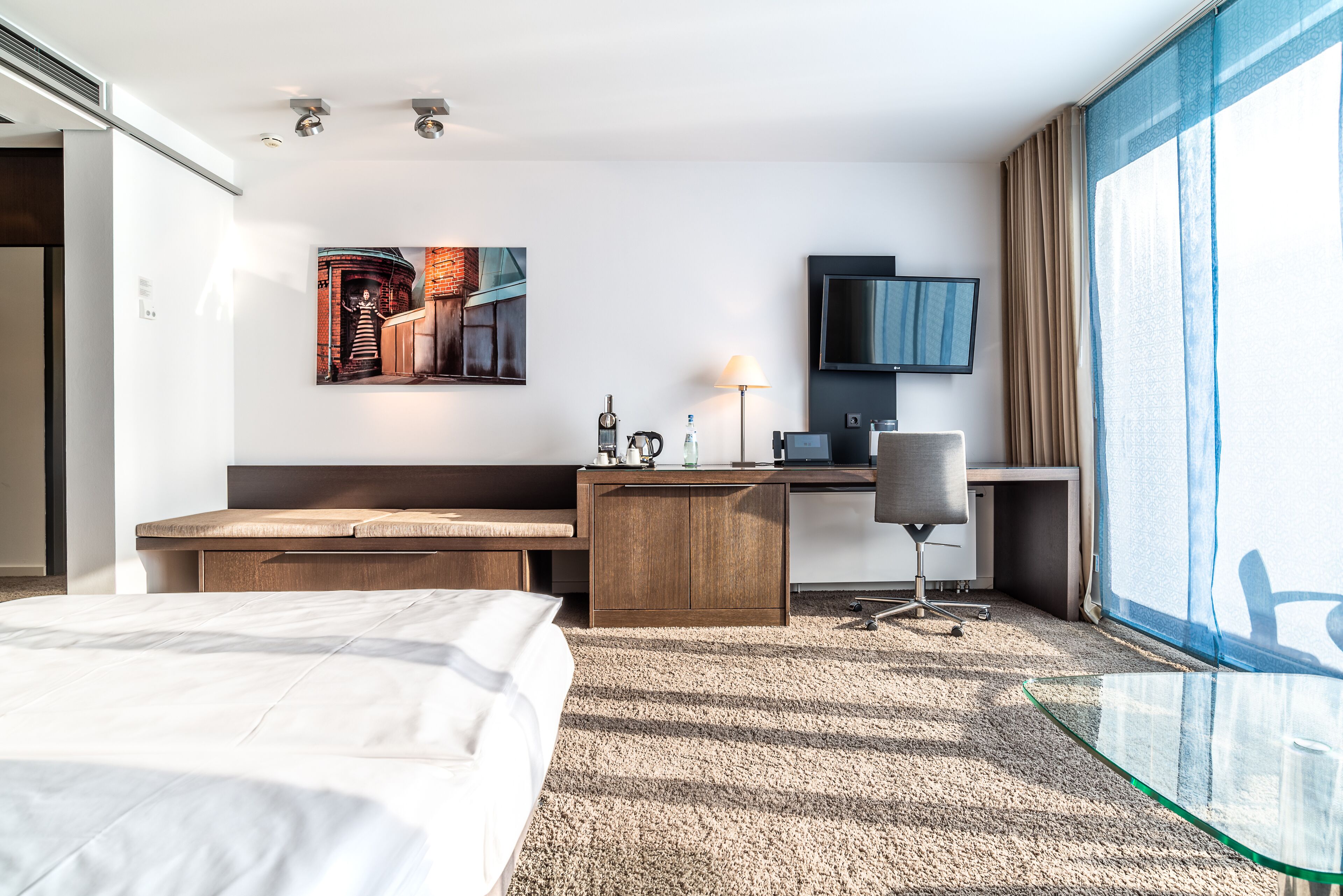 Habitación de hotel con cama, escritorio, silla, televisión y un ventanal con cortinas.