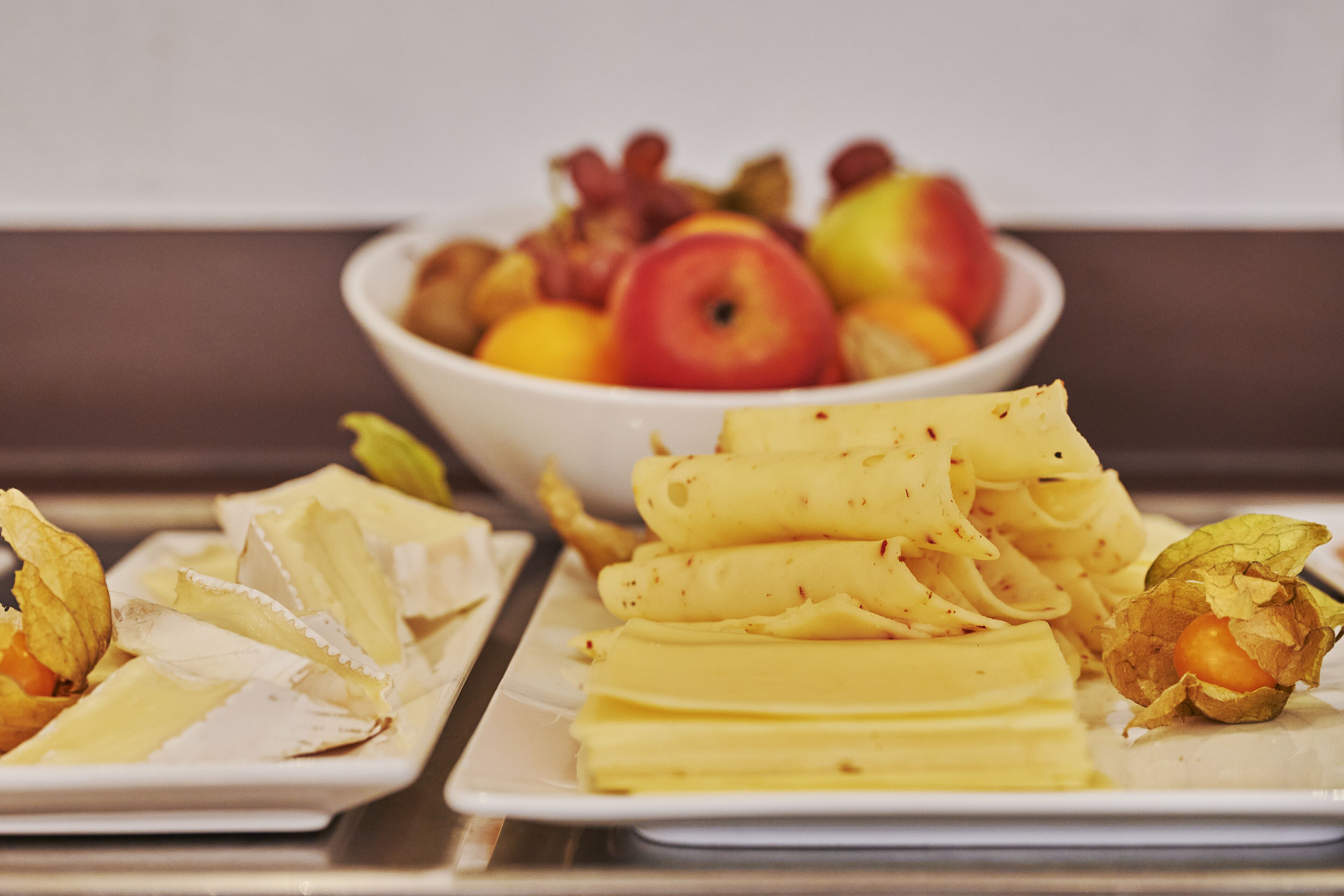 Un plato blanco con queso en rodajas y un bol de frutas variadas al fondo.