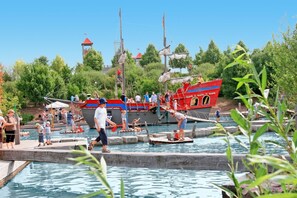 Um parque à beira-rio, com barcos, uma estrutura semelhante a um castelo e pessoas aproveitando a água.
