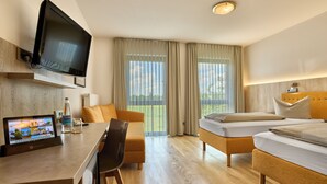 A hotel room with a flat-screen TV mounted above a wooden desk, a sofa, two single beds, and large windows with sheer curtains.