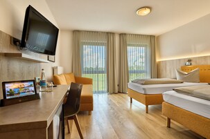 A hotel room with a flat-screen TV mounted above a wooden desk, a sofa, two single beds, and large windows with sheer curtains.