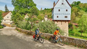 Two cyclists on a paved road with stone walls on one side and a large, white building with a red roof in the background.