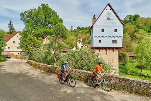 Two cyclists on a paved road with stone walls on one side and a large, white building with a red roof in the background.