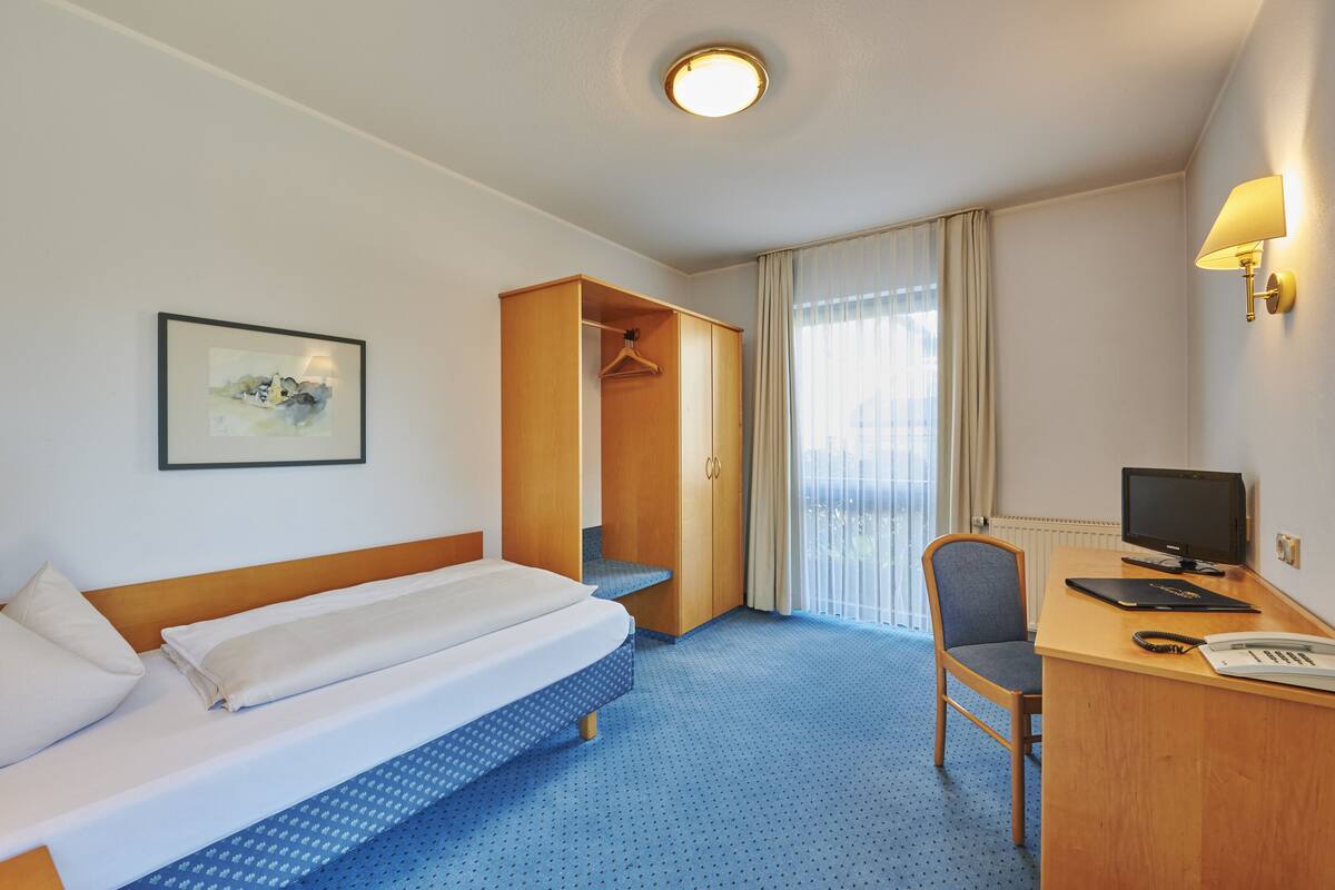 A hotel room with a bed, a desk with a computer, a chair, a coat rack, and a window with curtains.
