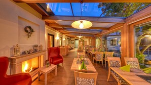 A cozy indoor dining area with wooden furniture, a fireplace, and a glass ceiling allowing natural light.