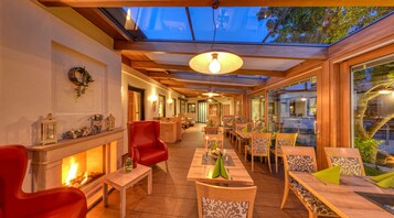 A cozy indoor dining area with wooden furniture, a fireplace, and a glass ceiling allowing natural light.