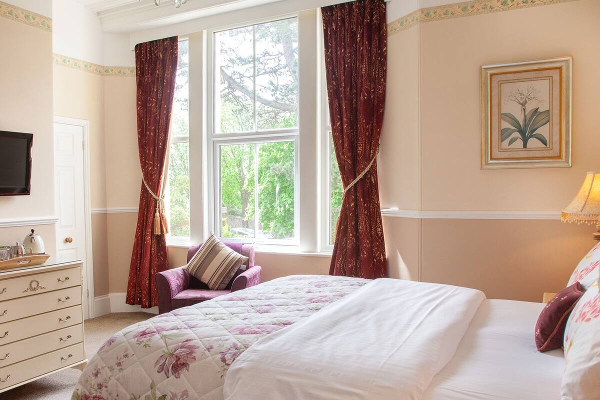 Ein Schlafzimmer mit einem Bett, einem Schminktisch, einem Sessel, einem Fenster mit Vorhängen und einem an der Wand montierten Fernseher.
