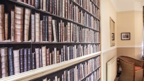 A library with tall bookshelves filled with books, a wicker chair, and a framed picture on the wall. - Court Prior Boutique B&B and Apartment (Torquay)