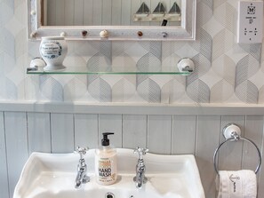 A bathroom with a white sink, two faucets, a glass shelf with decorative items, a mirror, and a towel ring.