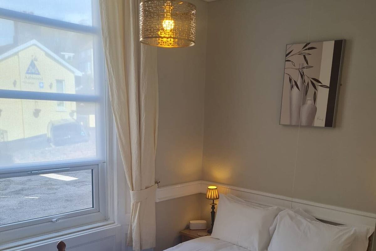 A neatly made bed with white linens and towels, a bedside lamp, and a window with curtains.