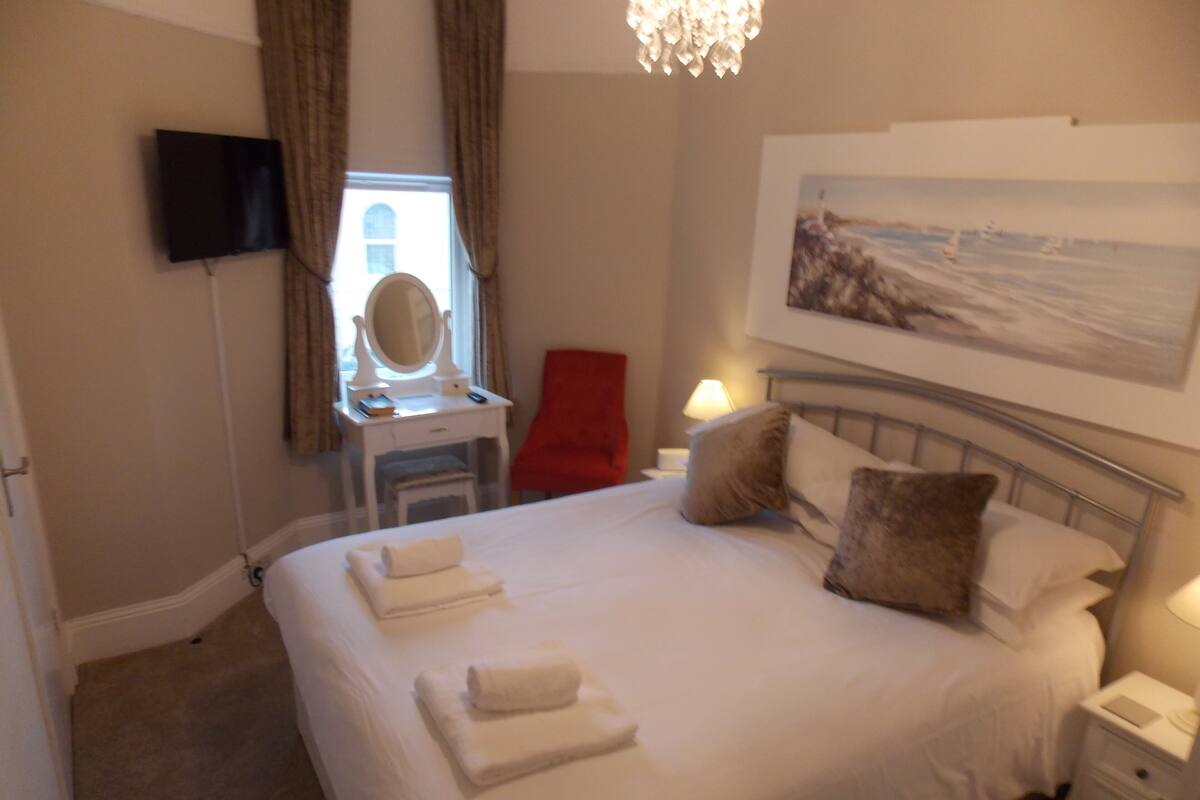 A hotel room with a bed, a desk with a mirror, a red chair, a nightstand with a lamp, and a painting on the wall.