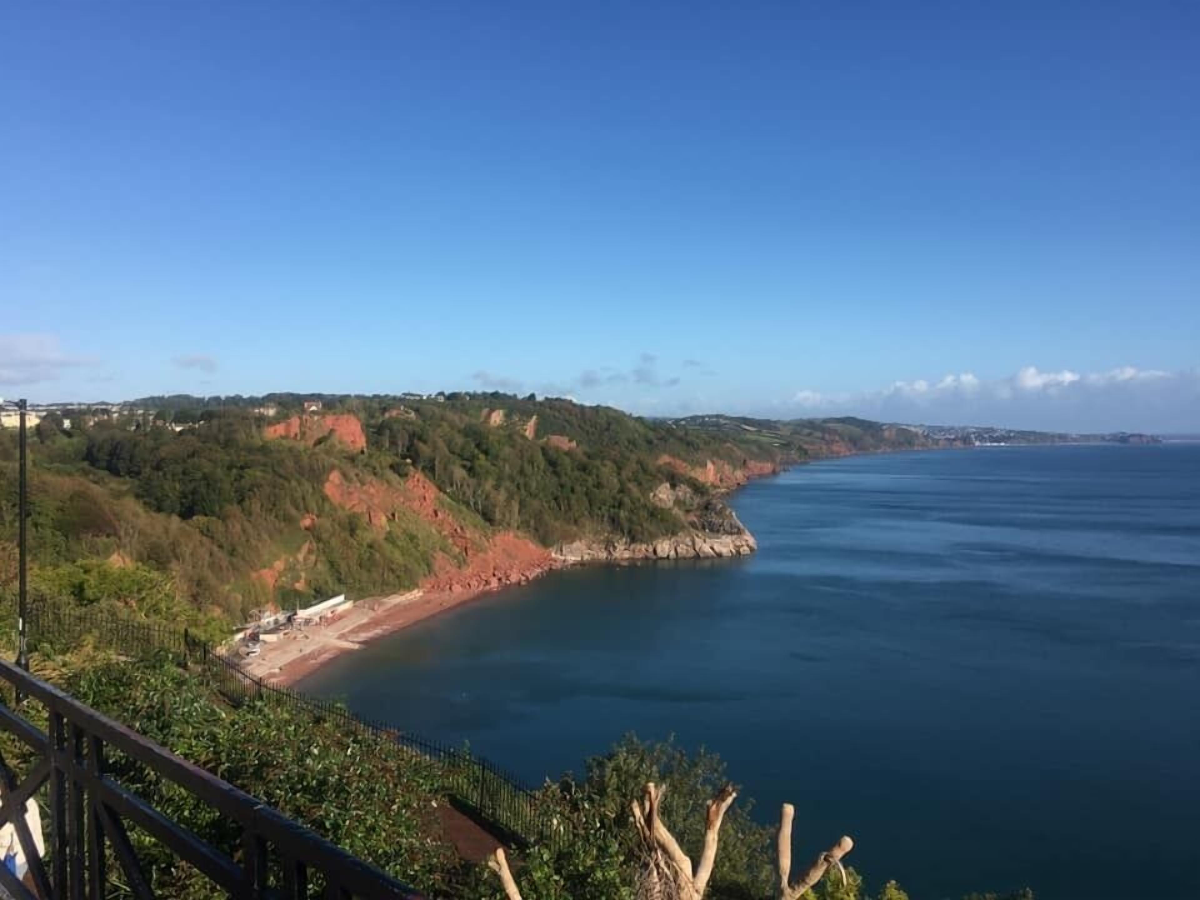 A coastal landscape with a clear view of the sea, a sandy beach, and a forested hillside.