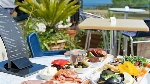 Una mesa servida con diversos platos, incluyendo mariscos, ensaladas y pan, al aire libre con vista al mar.