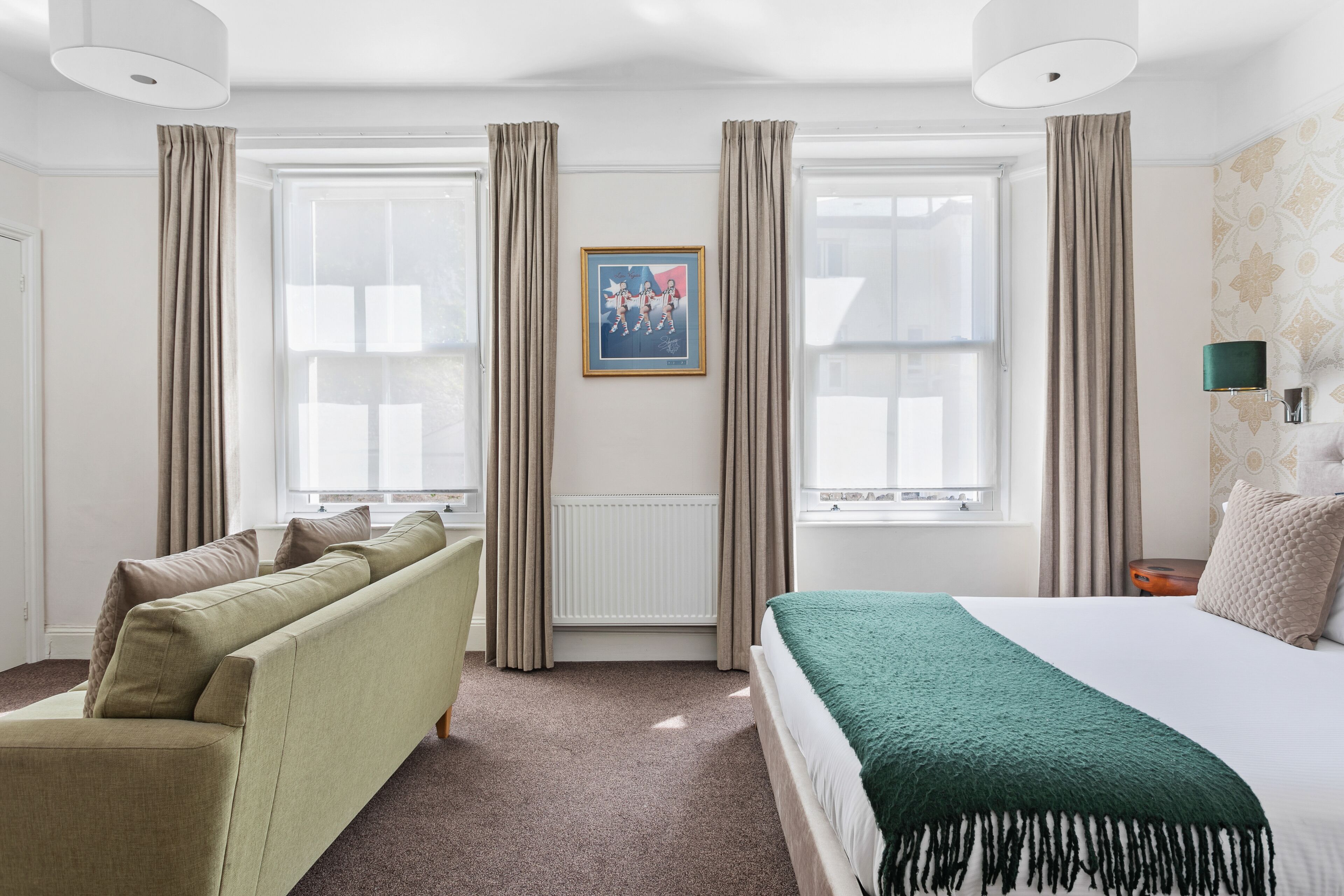 Habitación de hotel con cama, sofá, ventana con cortinas, radiador y un cuadro en la pared.