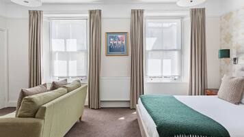 Habitación de hotel con cama, sofá, ventana con cortinas, radiador y un cuadro en la pared.