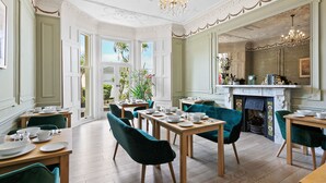 A dining area with wooden tables and green upholstered chairs, a chandelier, and a view of the garden through large windows. - The Torcroft (Torquay)