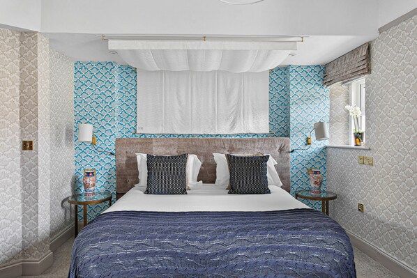 A bedroom with a large bed, two bedside tables, a nightstand, a mirror, and patterned wallpaper. - The Torcroft (Torquay)