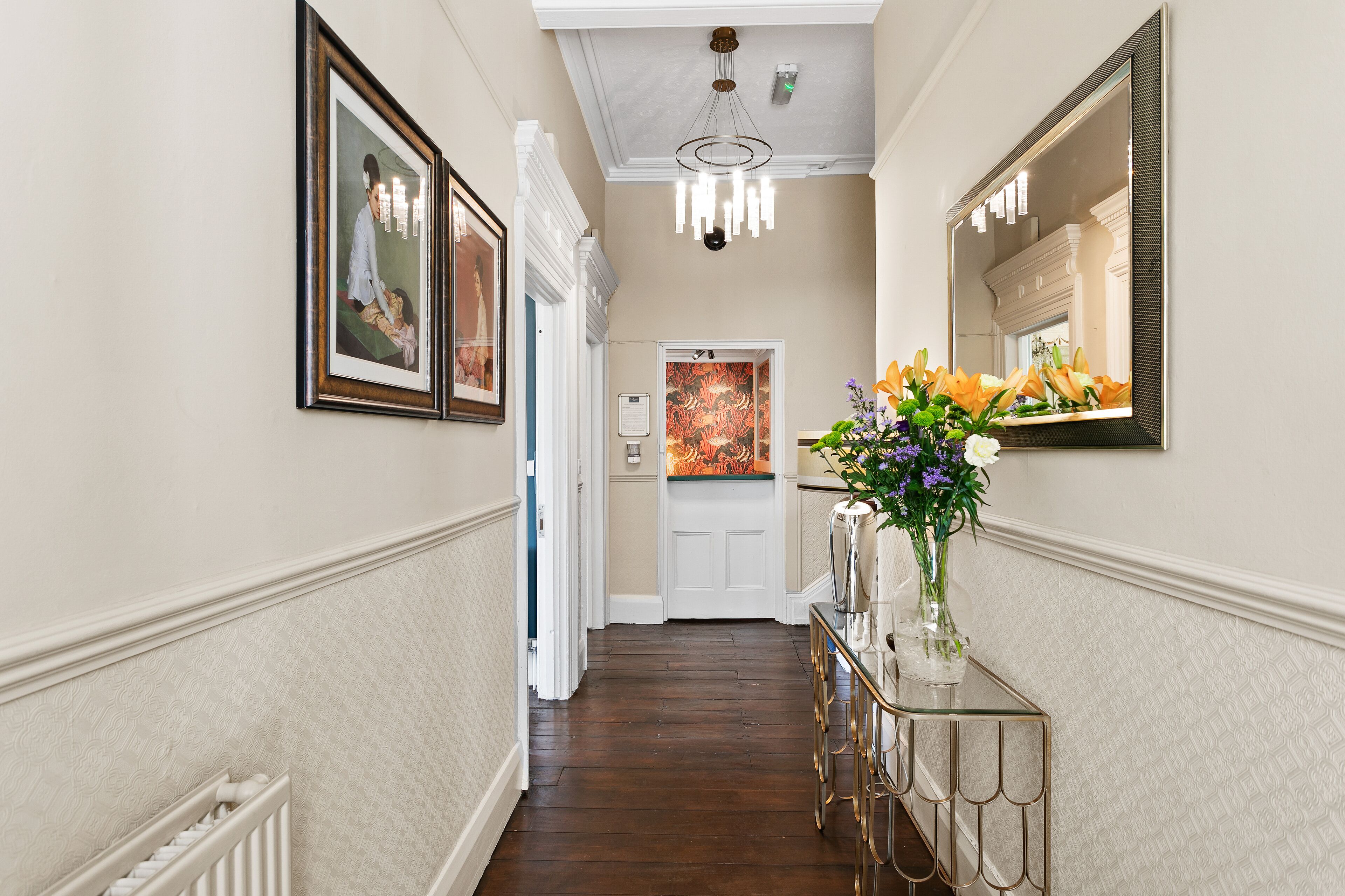 A hallway with framed artwork, a glass console table with a vase of flowers, and a door with a colorful painting.