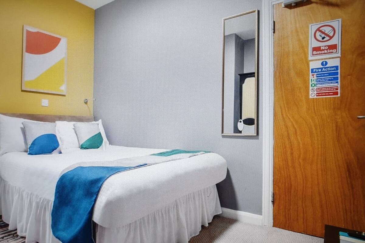 A hotel room with a bed, a nightstand, a mirror, and a door with a 'No Smoking' sign.