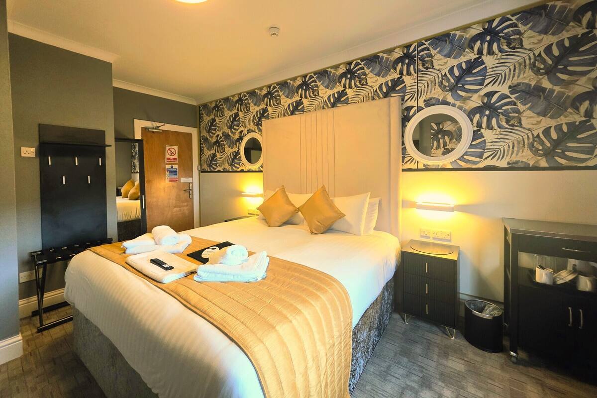 A hotel room with a large bed, a headboard with a mirror, a nightstand, a wardrobe, and a wall with a tropical leaf pattern.