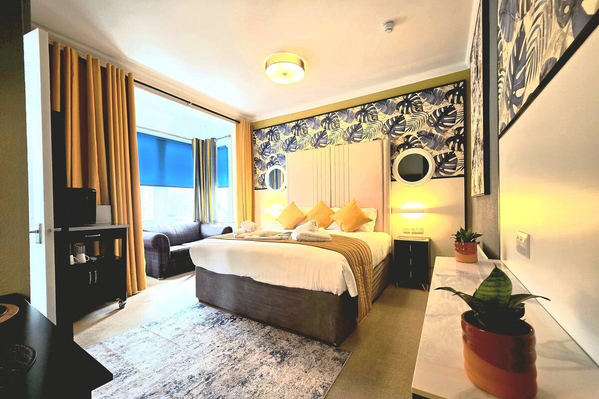A hotel room with a bed, bedside tables, a desk, a chair, a potted plant, and a window with curtains.