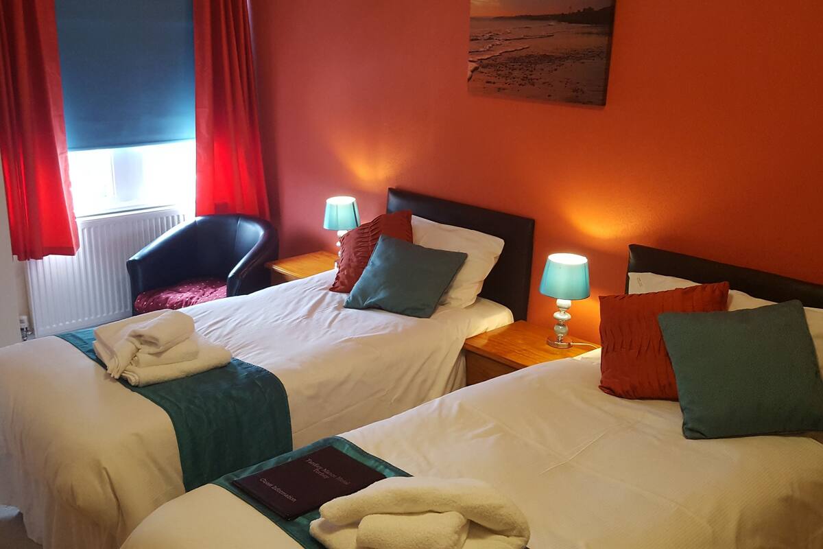 A hotel room with two beds, red curtains, a painting on the wall, and a view of the sea.