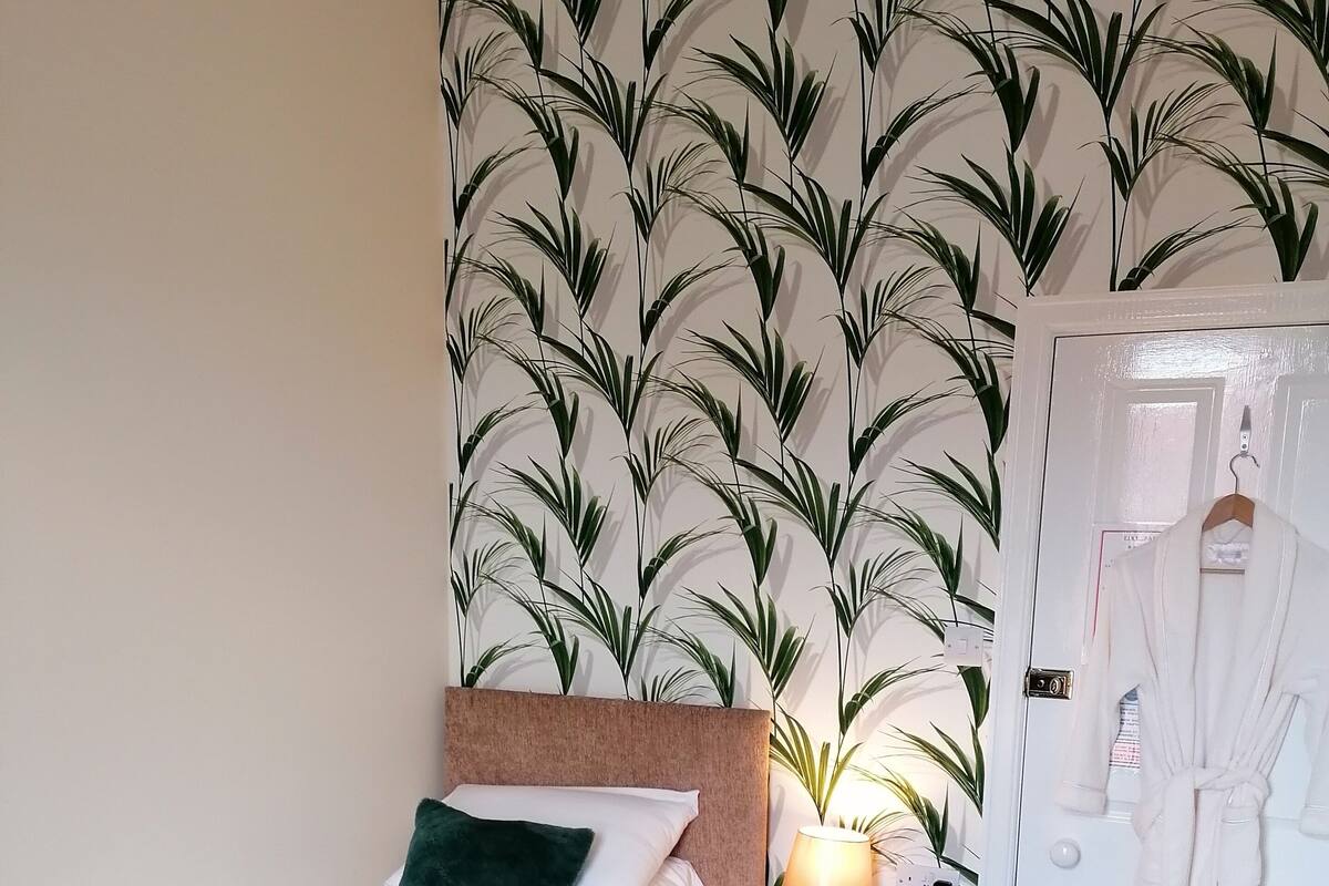 Quarto de hotel com cama, mesa de cabeceira, luminária, roupão pendurado na porta e papel de parede estampado.