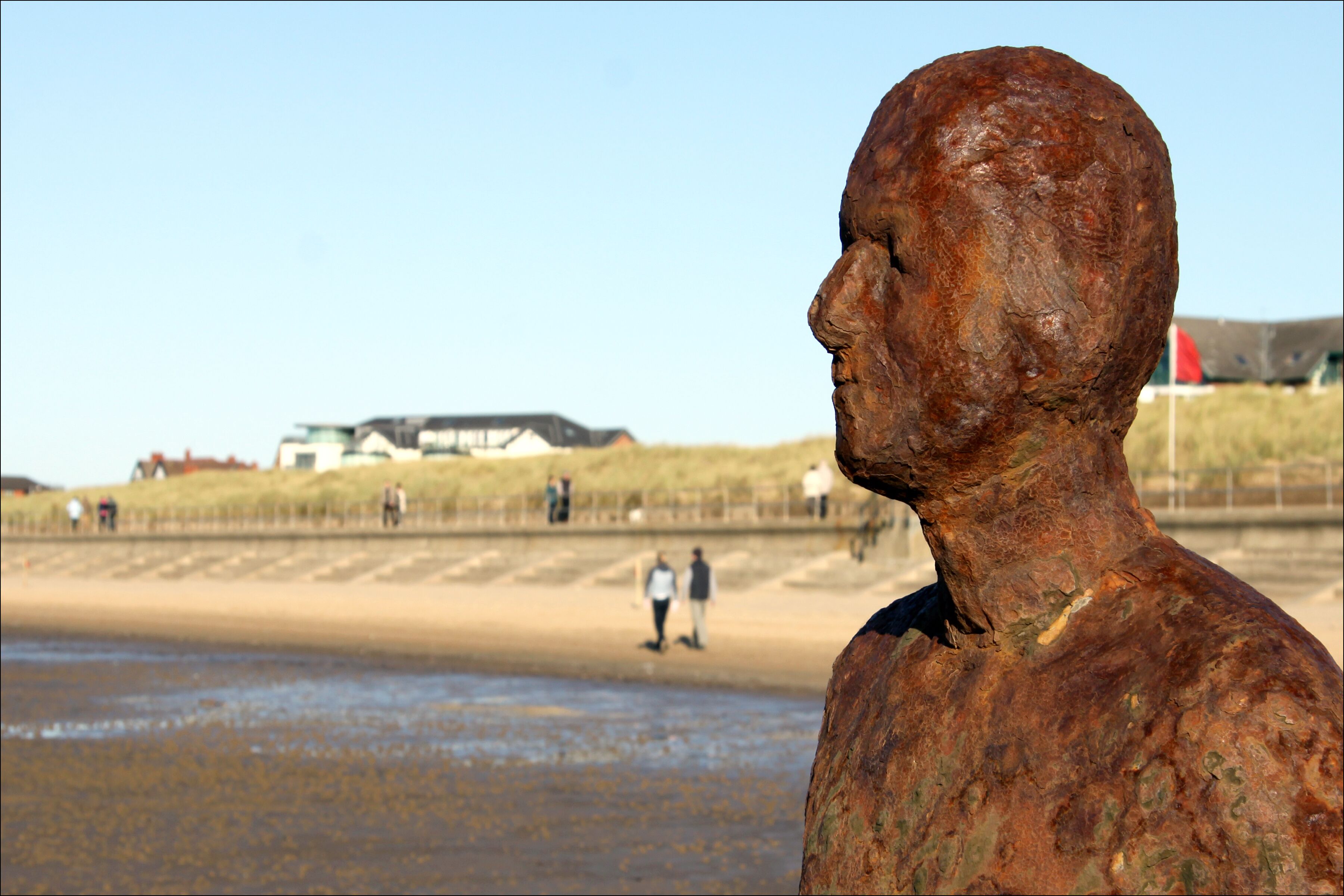 Una estatua de una figura humana en una playa, con gente de fondo.