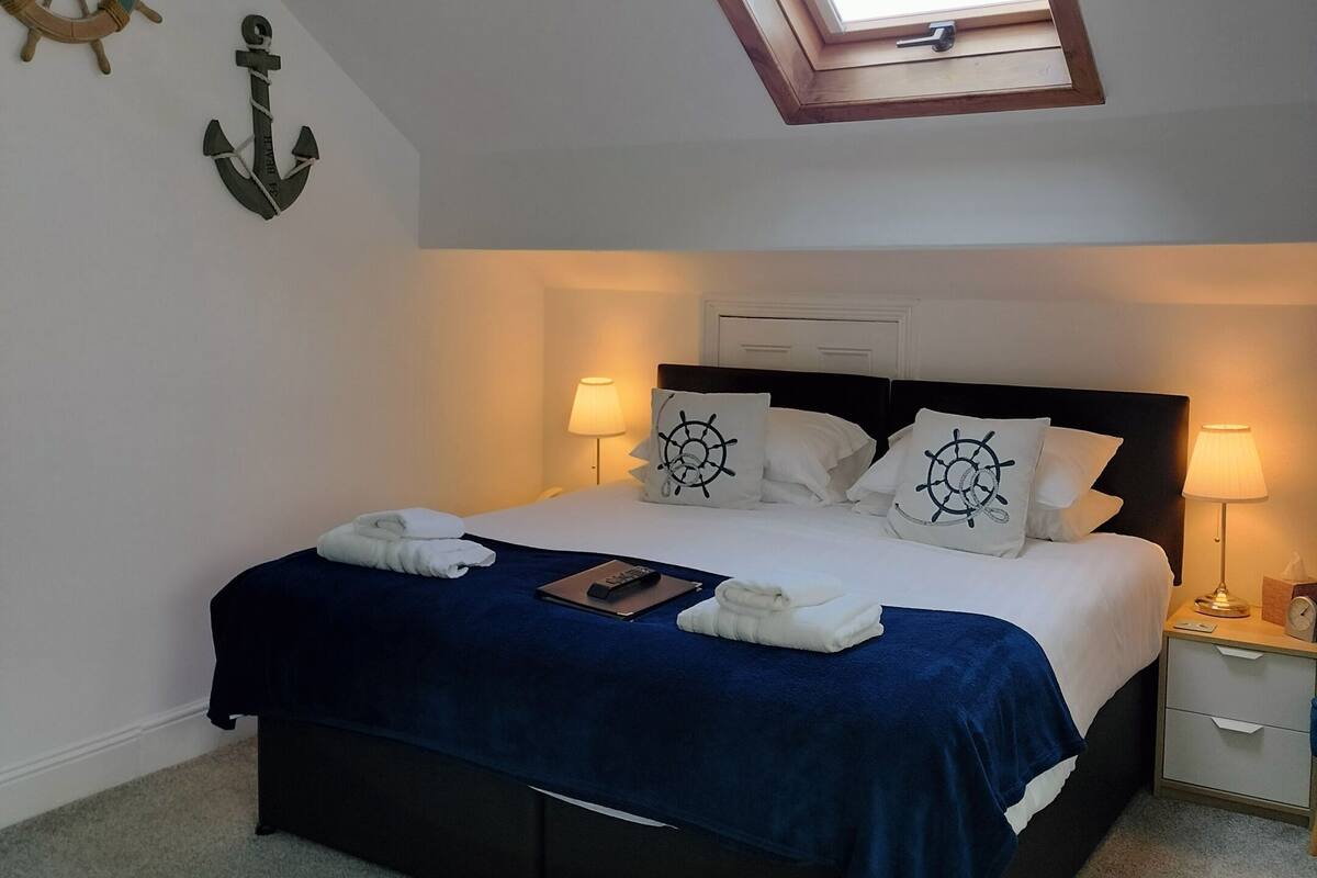 Cama bem arrumada com manta azul, duas luminárias de cabeceira, uma mesinha de cabeceira com relógio e um claraboia acima.