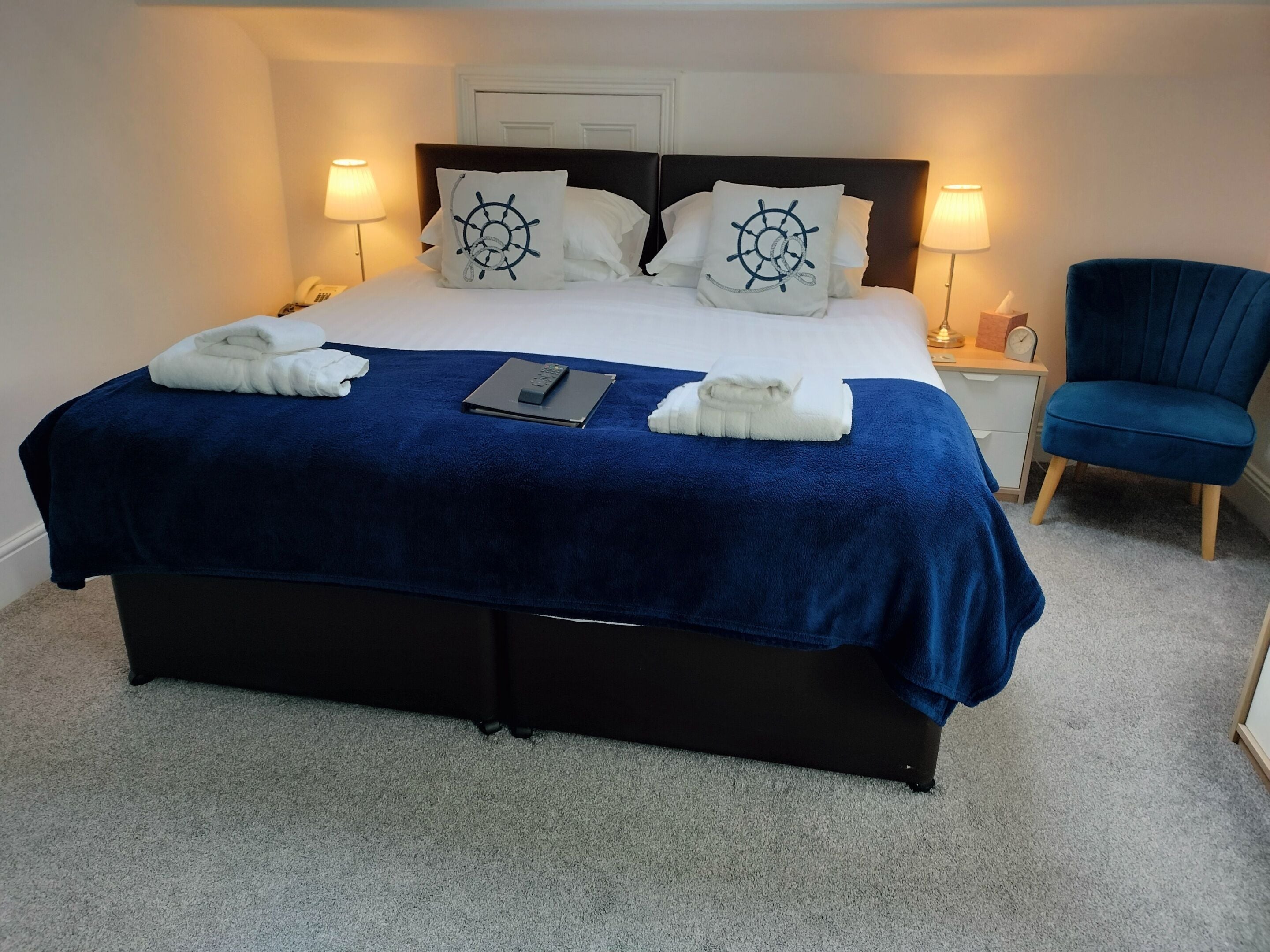 A neatly made bed with a blue bedspread, two white pillows with a ship wheel design, a remote control, and folded towels on the bed.