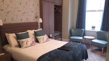 A hotel room with a bed, a wardrobe, a seating area with two chairs and a small table, and a window with curtains.
