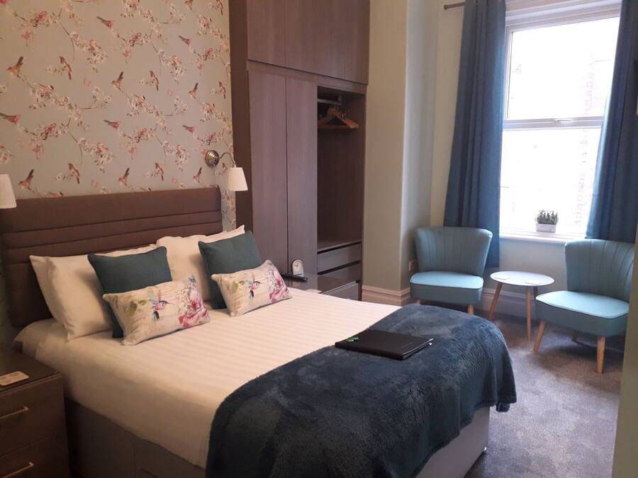 A hotel room with a bed, a wardrobe, a seating area with two chairs and a small table, and a window with curtains.