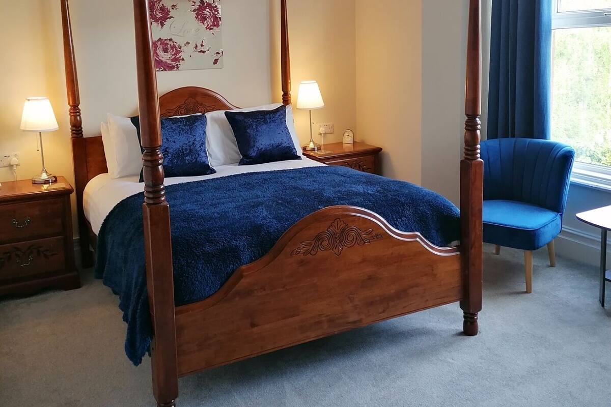 Uma cama de dossel com colcha e almofadas azuis, com mesas de cabeceira laterais com abajures, em um quarto com vista para o exterior através de uma janela com cortinas azuis.