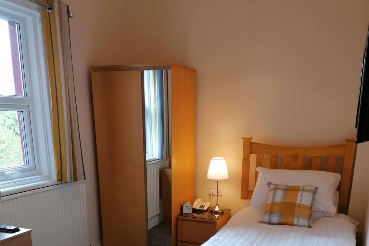 Quarto de hotel com cama, mesa de cabeceira com luminária, cadeira, escrivaninha e janela com cortinas.