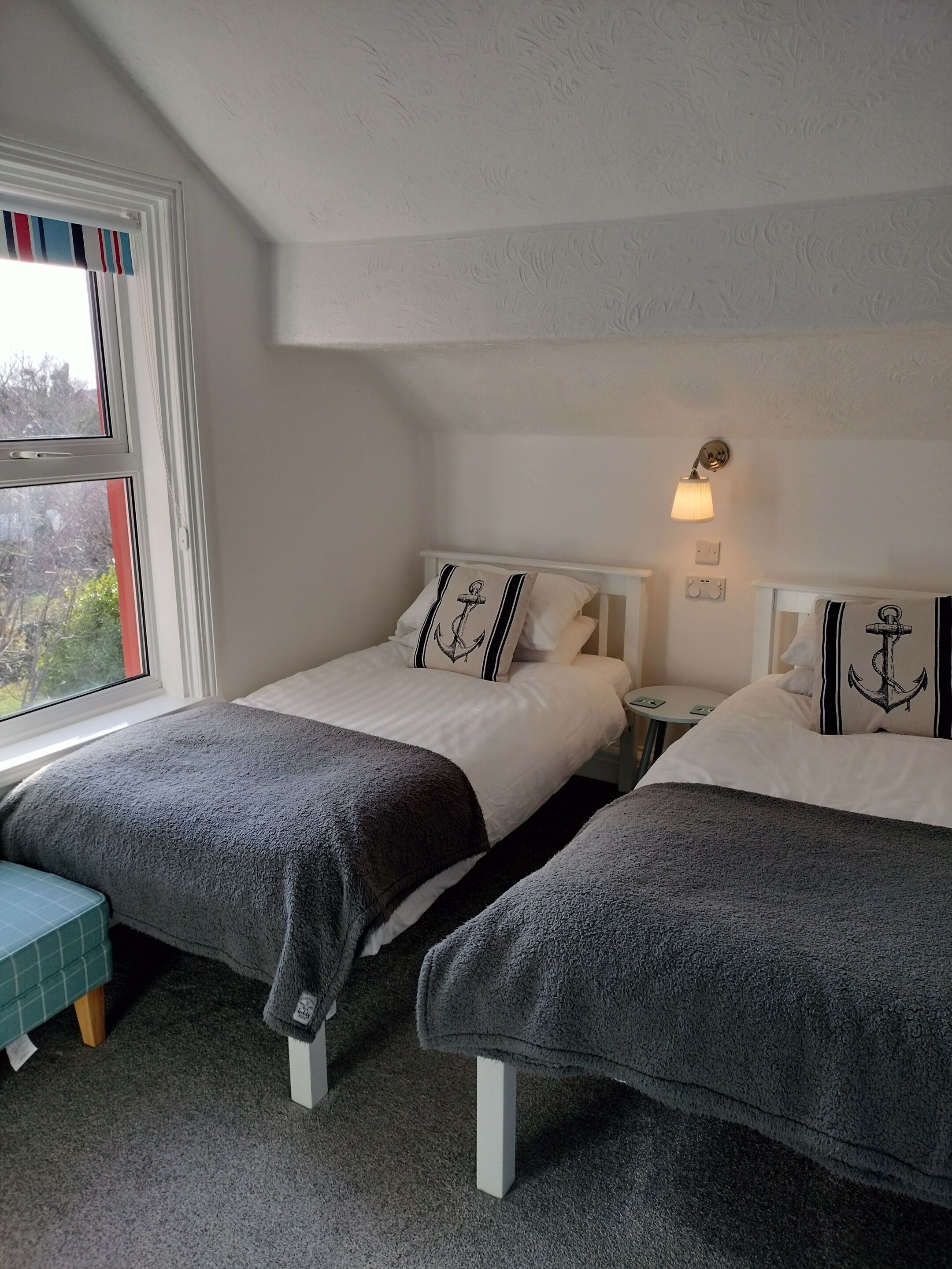 Habitación con dos camas, cada una con una manta gris y almohadas con diseño de ancla. Hay una ventana con cortinas a rayas, una mesita de noche con lámpara y una pequeña mesa redonda con una silla.