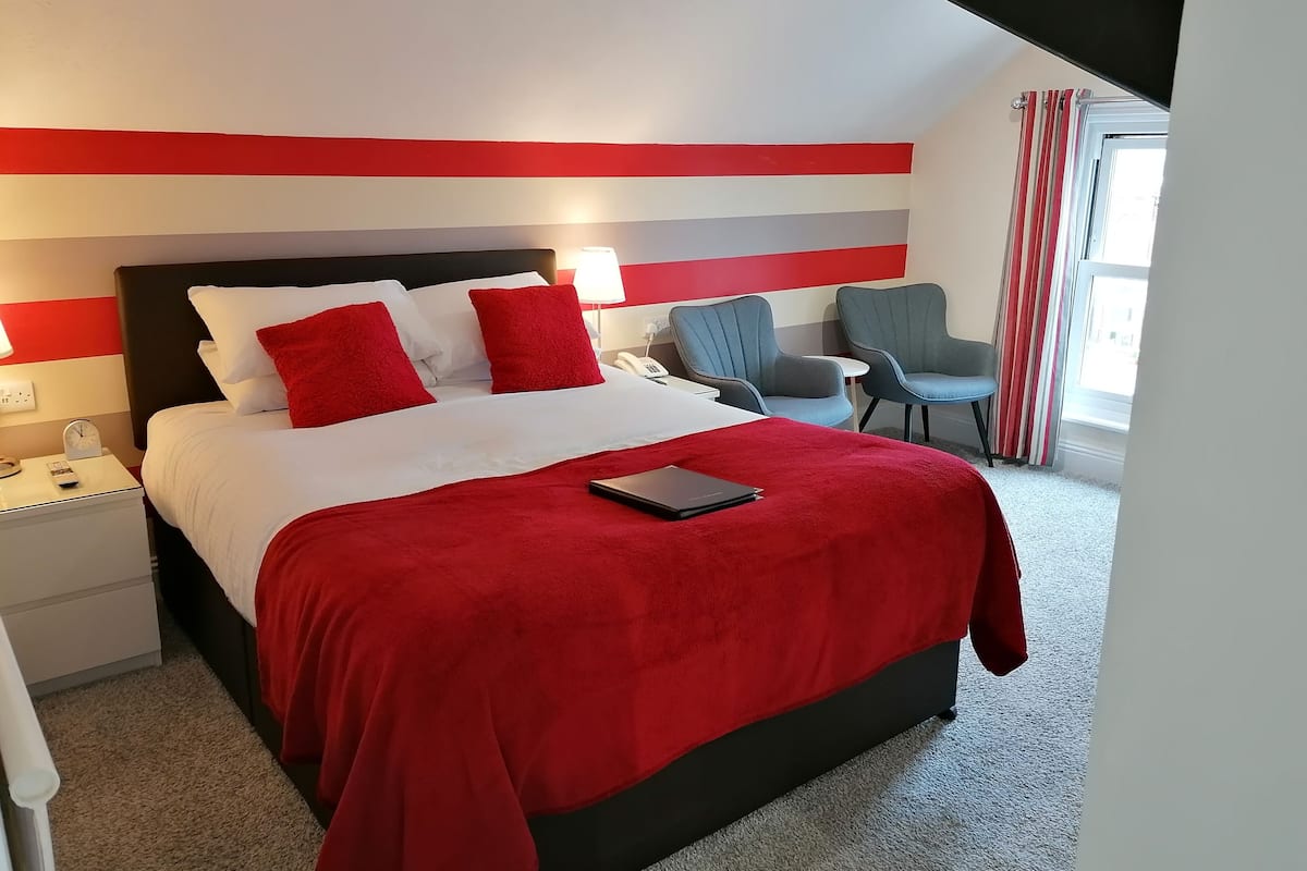 Quarto de hotel com cama, mesa de cabeceira, escrivaninha, duas cadeiras e janela com cortinas.