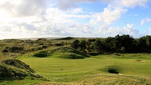 Un campo de golf con colinas onduladas, cielo despejado y vista distante del mar.